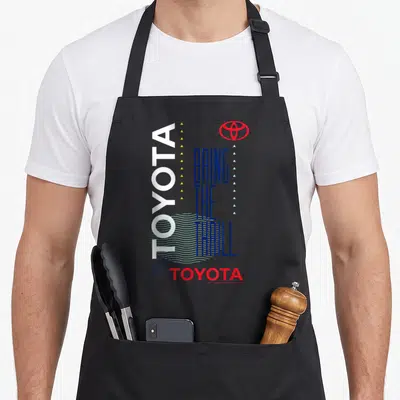 Bring toyota guy apron - Vertical white TOYOTA text, stacked blue slogan, red logo, yellow and light blue triangles — shown on a black cotton canvas apron against a white background