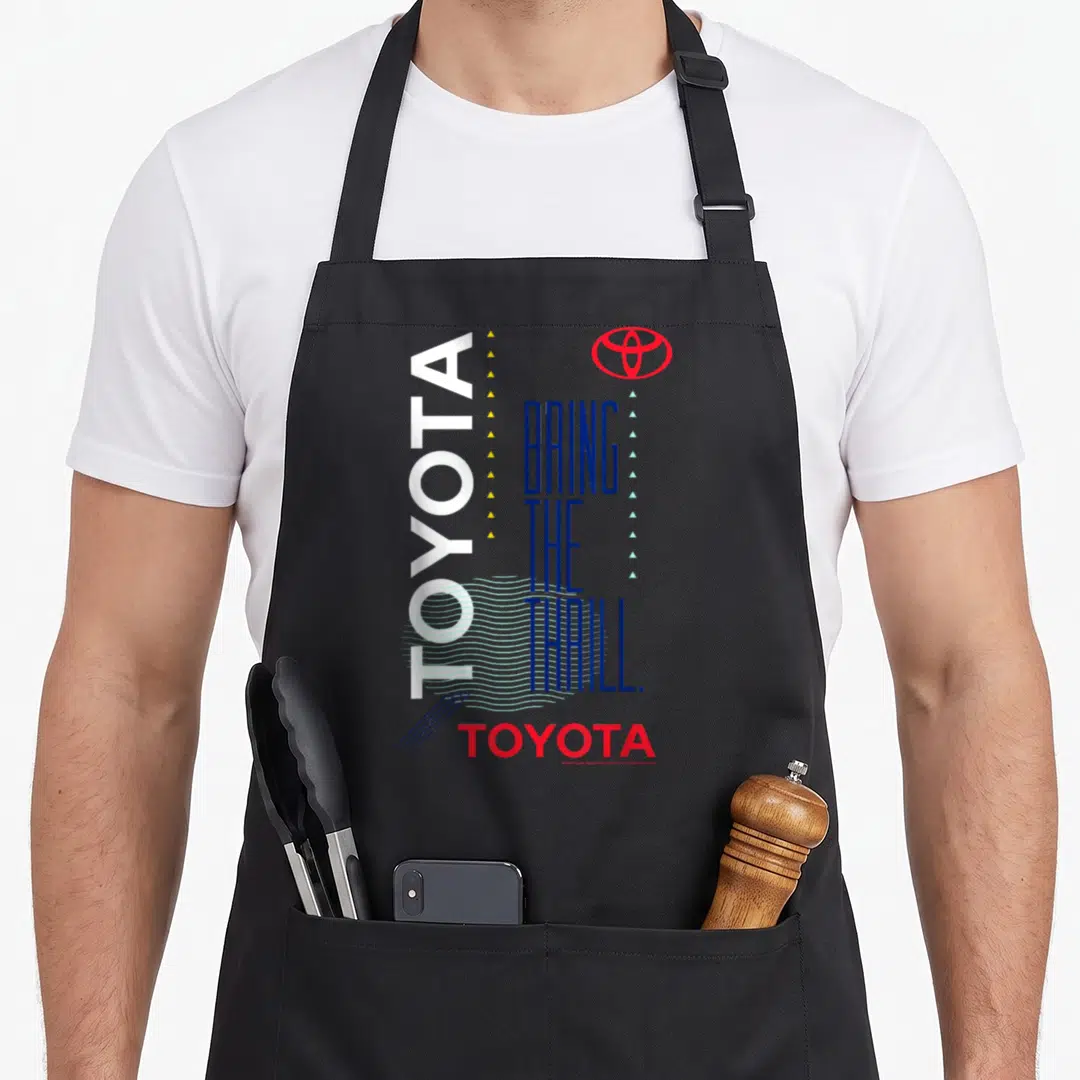 Bring toyota guy apron - Vertical white TOYOTA text, stacked blue slogan, red logo, yellow and light blue triangles — shown on a black cotton canvas apron against a white background