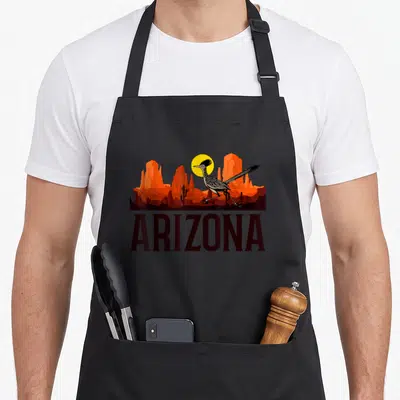 Arizona bird apron - Minimalist roadrunner running before orange canyons and yellow sun above bold ARIZONA text — shown on a black cotton canvas apron against a white background