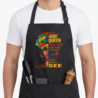 Quite queen apron - Woman headwrap I'M AN AUGUST QUEEN 3 sides text stars yellow red blue green brown — shown on a black cotton canvas apron against a white background