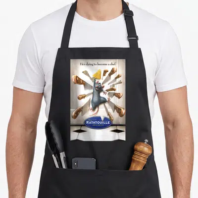 Hes ratatouille apron - Rat surrounded by flying knives and cheese with He's dying to become a chef text — shown on a black cotton canvas apron against a white background