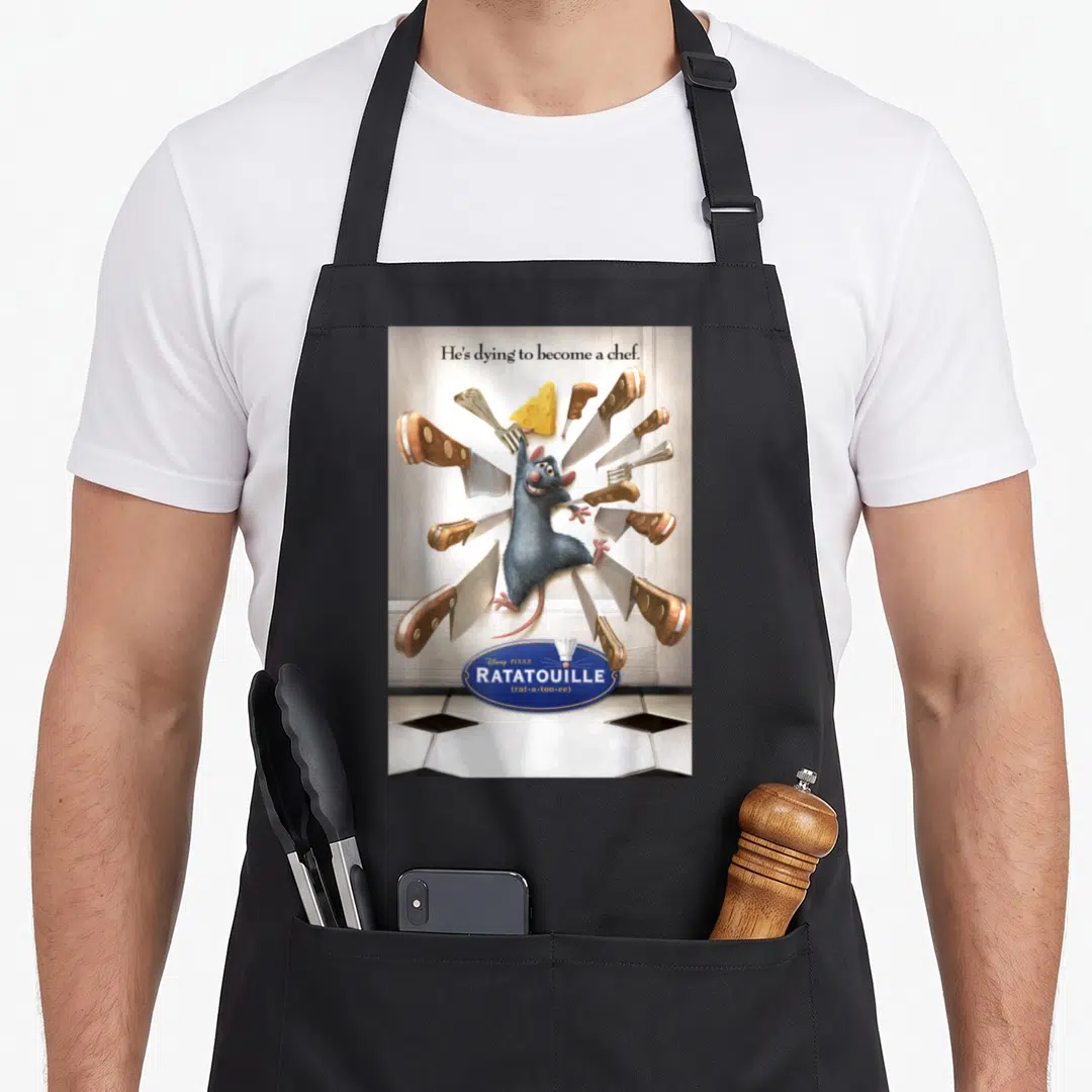 Hes ratatouille apron - Rat surrounded by flying knives and cheese with He's dying to become a chef text — shown on a black cotton canvas apron against a white background