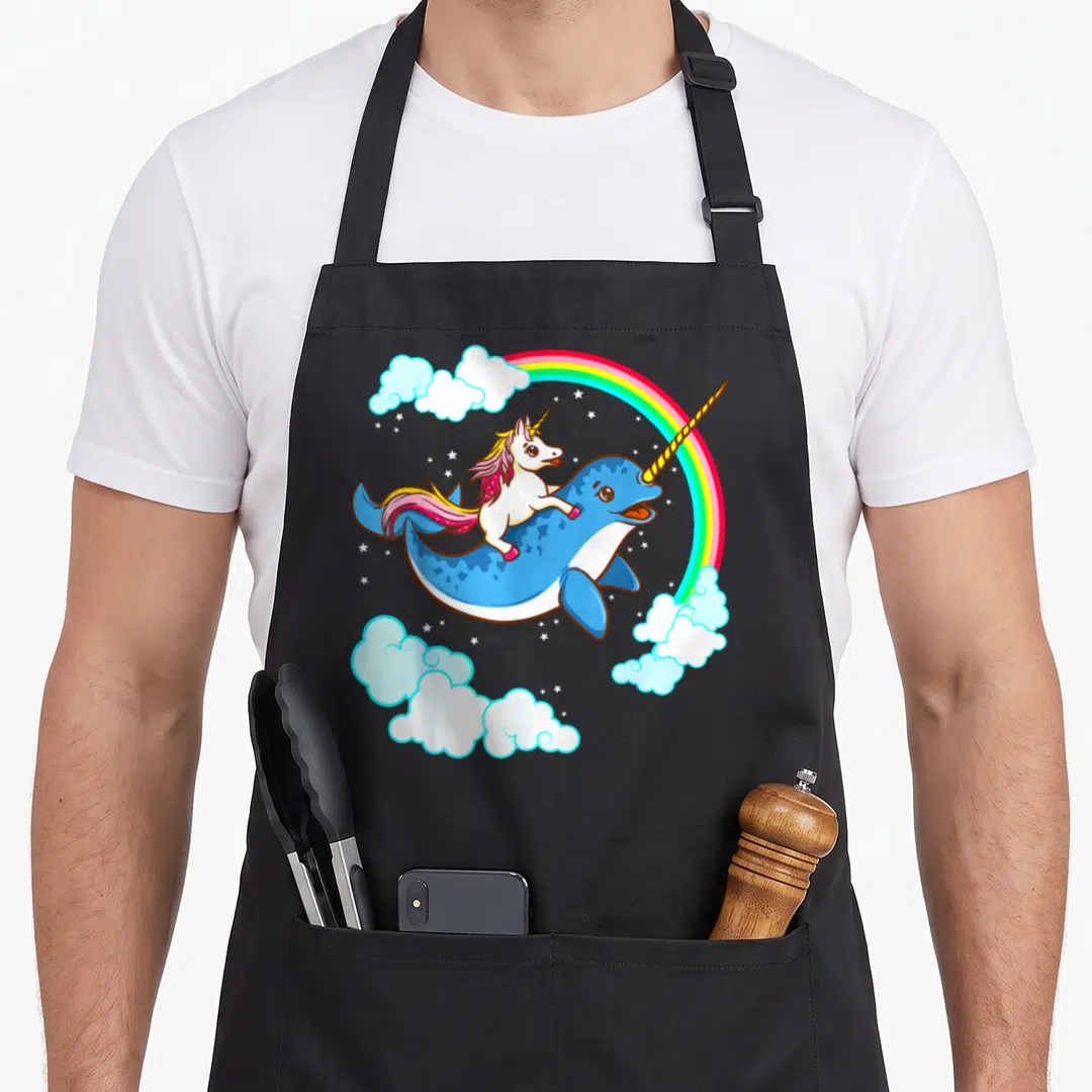 Narwhal youth unicorn apron - Cartoon unicorn riding blue narwhal under rainbow with clouds and stars in pink yellow — shown on a black cotton canvas apron against a white background