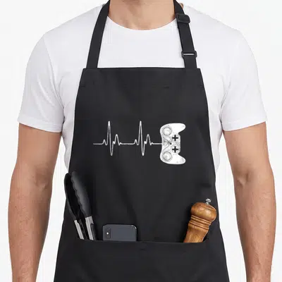 Cool gamer video game apron - White distressed EKG line connecting to a game controller graphic — shown on a black cotton canvas apron against a white background