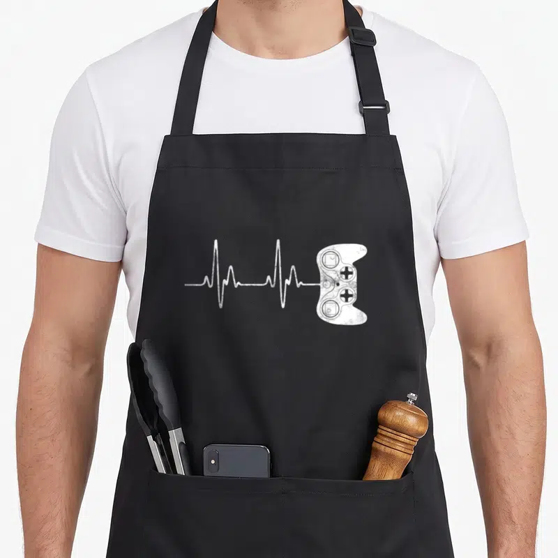 Cool gamer video game apron - White distressed EKG line connecting to a game controller graphic — shown on a black cotton canvas apron against a white background