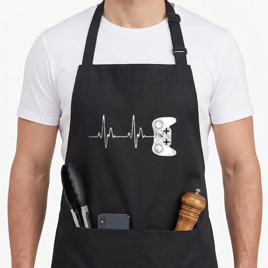 Cool gamer video game apron - White distressed EKG line connecting to a game controller graphic — shown on a black cotton canvas apron against a white background