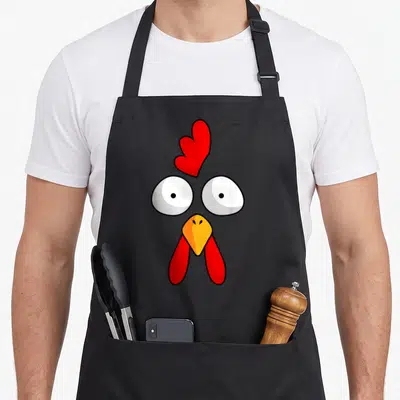 Face rooster apron - Cartoon chicken face with red comb and wattle, yellow beak, and white eyes on grey background — shown on a black cotton canvas apron against a white background