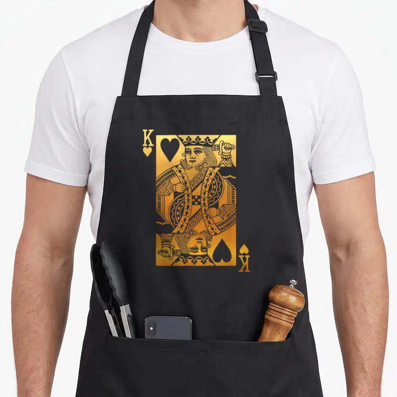 Card king apron - Gold mirrored king playing card with K and heart symbols in corners — shown on a black cotton canvas apron against a white background