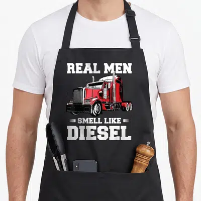 Husband semi truck apron - Red semi-truck illustration with Real Men Smell Like Diesel text — shown on a black cotton canvas apron against a white background