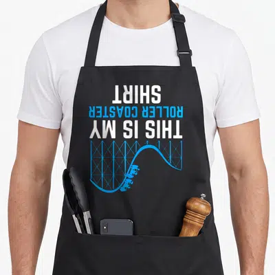 Funny coaster roller apron - Upside down text reading THIS IS MY ROLLER COASTER SHIRT above blue track — shown on a black cotton canvas apron against a white background