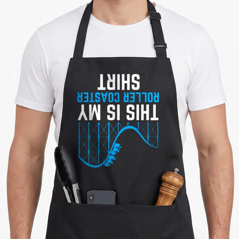 Funny coaster roller apron - Upside down text reading THIS IS MY ROLLER COASTER SHIRT above blue track — shown on a black cotton canvas apron against a white background