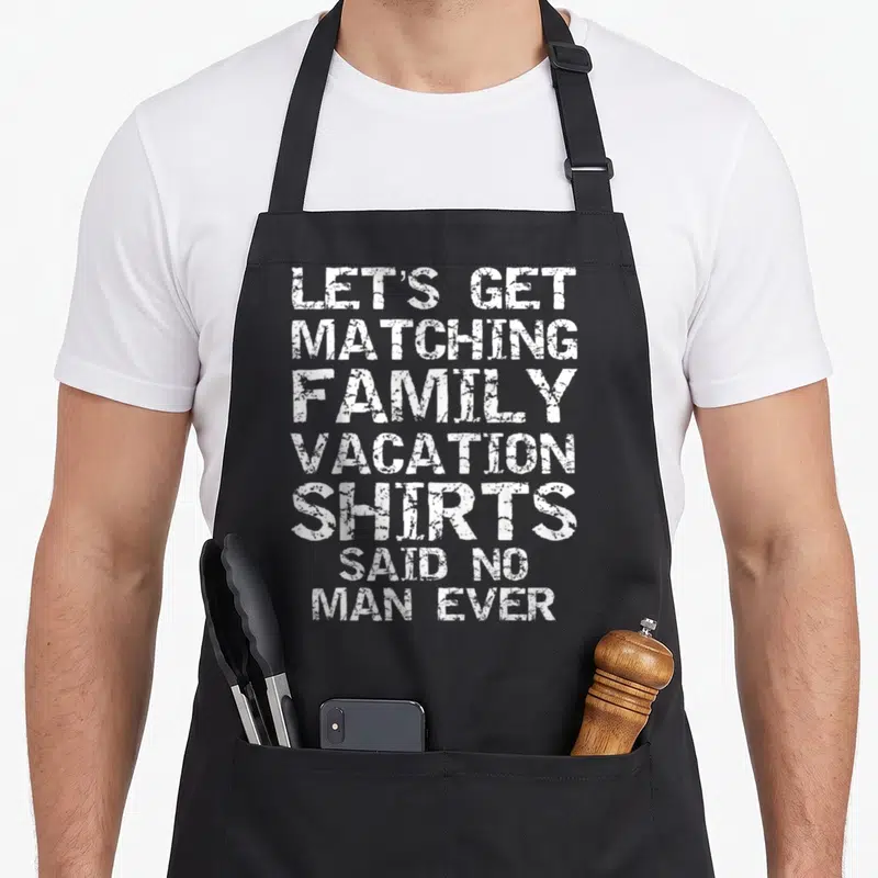 Funny vacation family aprons - White stacked text reading LET'S GET MATCHING FAMILY VACATION SHIRTS SAID NO MAN EVER — shown on a black cotton canvas apron against a white background