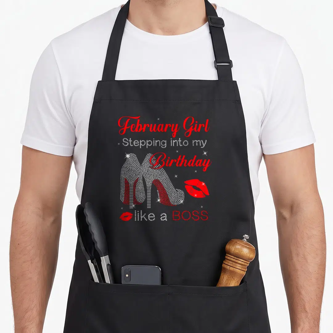 Birthday bbq boss apron - Rhinestone high heels and red lips with stacked text reading February Girl Stepping into my — shown on a black cotton canvas apron against a white background