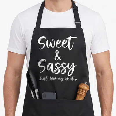 Sweet aunt sassy apron - White stacked text reading Sweet Sassy Just like my aunt with a small heart icon — shown on a black cotton canvas apron against a white background