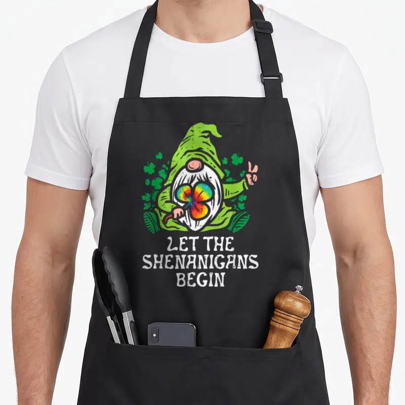 Gnome st patrick day aprons - Cartoon gnome holding rainbow shamrock above LET THE SHENANIGANS BEGIN text — shown on a black cotton canvas apron against a white background
