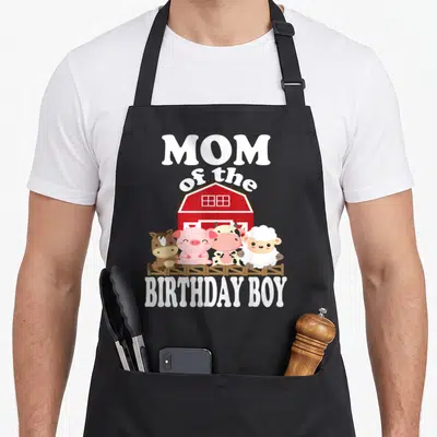 Birthday farming apron - MOM of the BIRTHDAY BOY text with four cartoon farm animals standing by a red barn — shown on a black cotton canvas apron against a white background