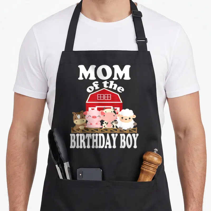 Birthday farming apron - MOM of the BIRTHDAY BOY text with four cartoon farm animals standing by a red barn — shown on a black cotton canvas apron against a white background