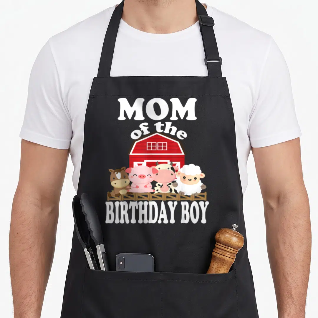 Birthday farming apron - MOM of the BIRTHDAY BOY text with four cartoon farm animals standing by a red barn — shown on a black cotton canvas apron against a white background