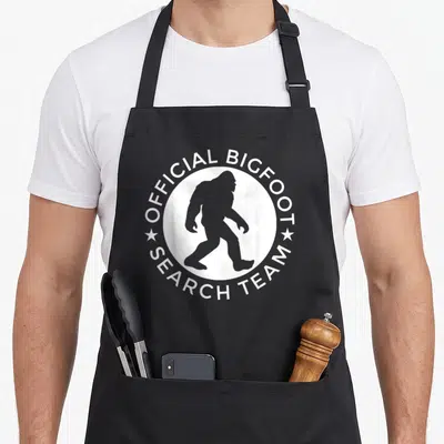 Search yeti apron - Grey Bigfoot silhouette in white circle with Official Bigfoot Search Team text — shown on a black cotton canvas apron against a white background