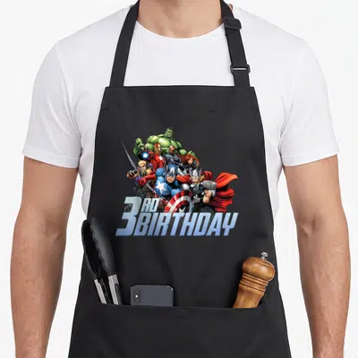 Shot 3rd birthday apron - Realistic Avengers group print with 3RD BIRTHDAY text in blue and red — shown on a black cotton canvas apron against a white background