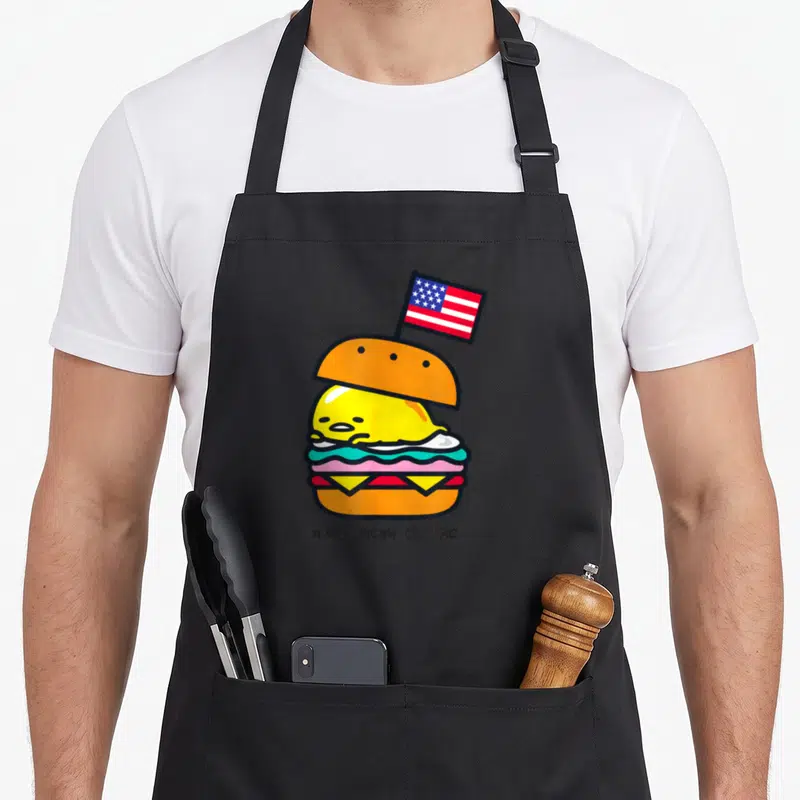 Burger gudetama apron - Cartoon egg burger with US flag and A-Meh-Rican Classic text — shown on a black cotton canvas apron against a white background