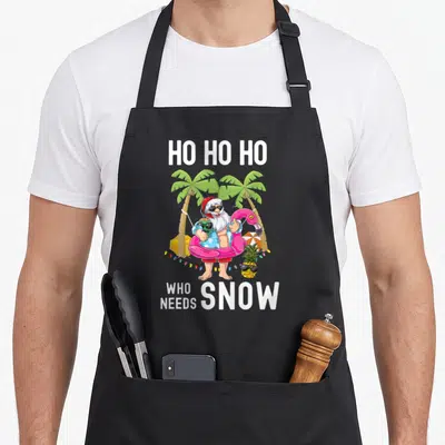 Surfing christmas tree apron - Santa in pink flamingo floatie with HO HO HO WHO NEEDS SNOW text and palm trees — shown on a black cotton canvas apron against a white background