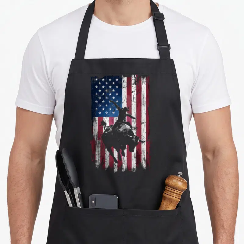 Flag western aprons - Grey bull rider silhouette on distressed red white blue USA flag — shown on a black cotton canvas apron against a white background