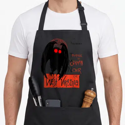 Mothman art class apron - Black Mothman figure with orange halo and French text Prochainement Tournée du Mothman Noir — shown on a black cotton canvas apron against a white background