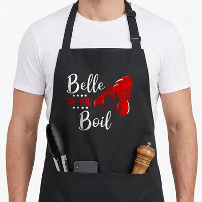 Belle seafood boil apron - Red cartoon lobster next to stacked white and red text separated by white dots — shown on a black cotton canvas apron against a white background