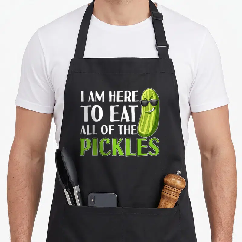 Funny eat pickle apron - Stacked text I AM HERE TO EAT ALL OF THE PICKLES next to a green cartoon pickle with black — shown on a black cotton canvas apron against a white background