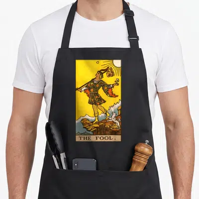 Fool major apron - Man standing on cliff with dog and THE FOOL text in retro yellow and green style — shown on a black cotton canvas apron against a white background