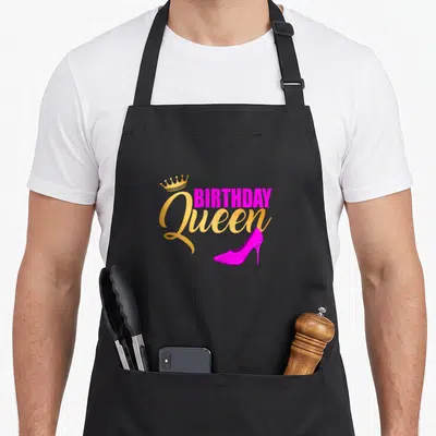 Bday queen of baking apron - Gold crown, magenta BIRTHDAY Queen text, and magenta high heel shoe graphic — shown on a black cotton canvas apron against a white background