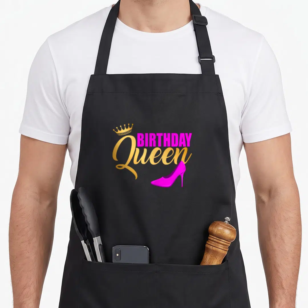 Bday queen of baking apron - Gold crown, magenta BIRTHDAY Queen text, and magenta high heel shoe graphic — shown on a black cotton canvas apron against a white background