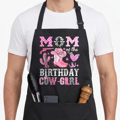 Cow cowgirl aprons - MOM of the BIRTHDAY COW-GIRL text surrounding pink boots, a cowboy hat, revolver, cacti — shown on a black cotton canvas apron against a white background