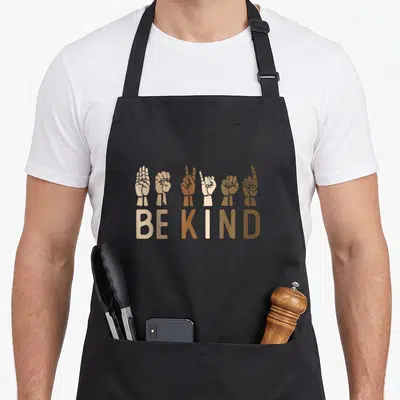 Kindness asl sign for apron - Six hands in varying skin tones forming ASL signs above multi-shade brown text reading BE — shown on a black cotton canvas apron against a white background