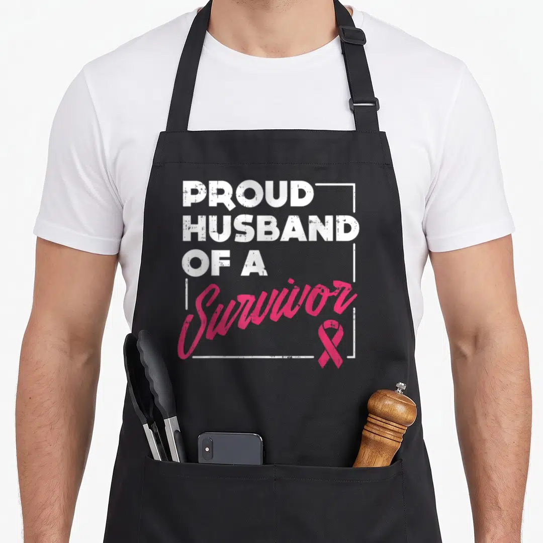 Proud breast cancer awareness aprons - Stacked white text reading Proud Husband of a Survivor with pink ribbon — shown on a black cotton canvas apron against a white background