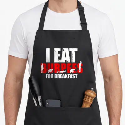 Eat burpees apron - I EAT BURPEES FOR BREAKFAST text with white exercise silhouettes inside red letters — shown on a black cotton canvas apron against a white background