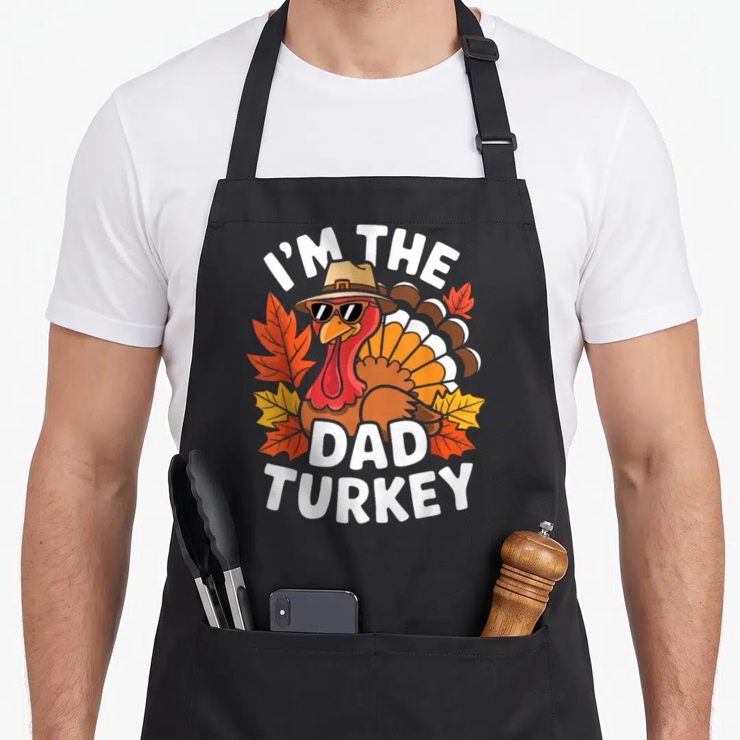 Family costume thanksgiving apron - Cartoon turkey wearing sunglasses and hat with I'M THE DAD TURKEY text surrounded — shown on a black cotton canvas apron against a white background