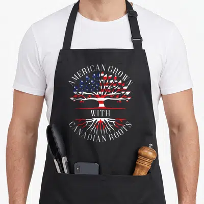 Canadian grown pride apron - Tree graphic with American flag leaves and Canadian flag roots under arched text — shown on a black cotton canvas apron against a white background