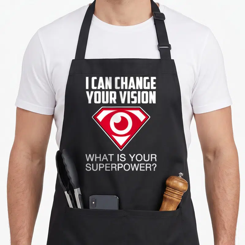Optometry doctor apron - I CAN CHANGE YOUR VISION text and red shield eye symbol on white background — shown on a black cotton canvas apron against a white background
