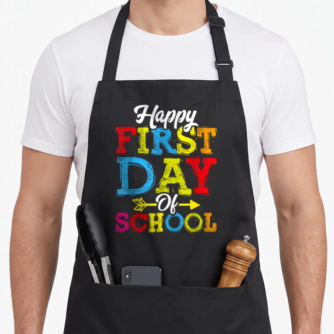 Cool school teacher apron - Stacked colorful text reading Happy First Day Of School with yellow arrow on black — shown on a black cotton canvas apron against a white background