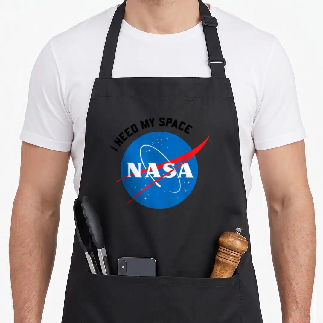 Funny space nasa apron - I NEED MY SPACE text over blue NASA circle with red swoosh — shown on a black cotton canvas apron against a white background