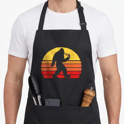Sasquatch pizza apron - Grey Bigfoot silhouette eating pepperoni pizza slice in front of retro yellow orange and red — shown on a black cotton canvas apron against a white background