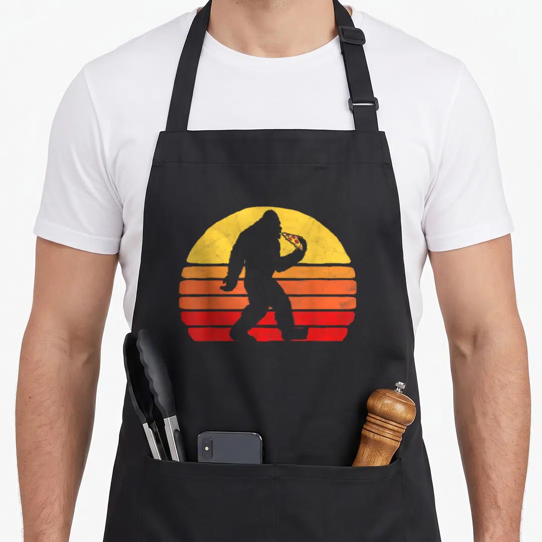 Sasquatch pizza apron - Grey Bigfoot silhouette eating pepperoni pizza slice in front of retro yellow orange and red — shown on a black cotton canvas apron against a white background