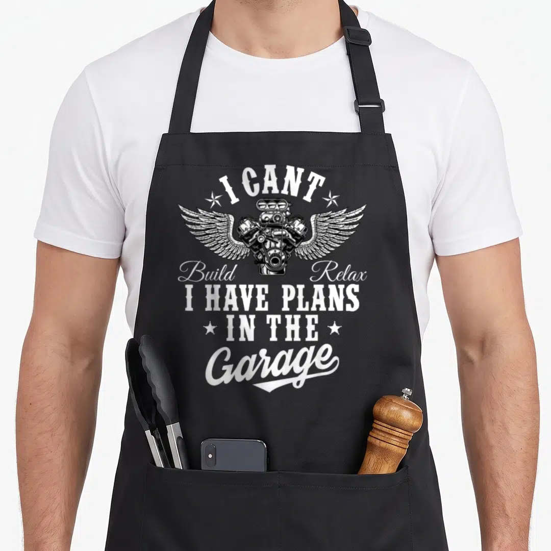Garage father day apron - Winged engine illustration surrounded by white text reading I cant build relax I have plans — shown on a black cotton canvas apron against a white background