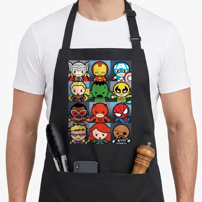 Boxed kawaii apron - Twelve superhero cartoon figures in red blue yellow green 4x3 grid squares — shown on a black cotton canvas apron against a white background