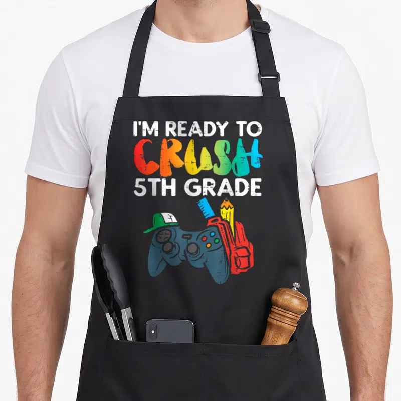 Crush ready grade apron - Rainbow text I'M READY TO CRUSH 5TH GRADE above game controller, red backpack, pencil, ruler — shown on a black cotton canvas apron against a white background