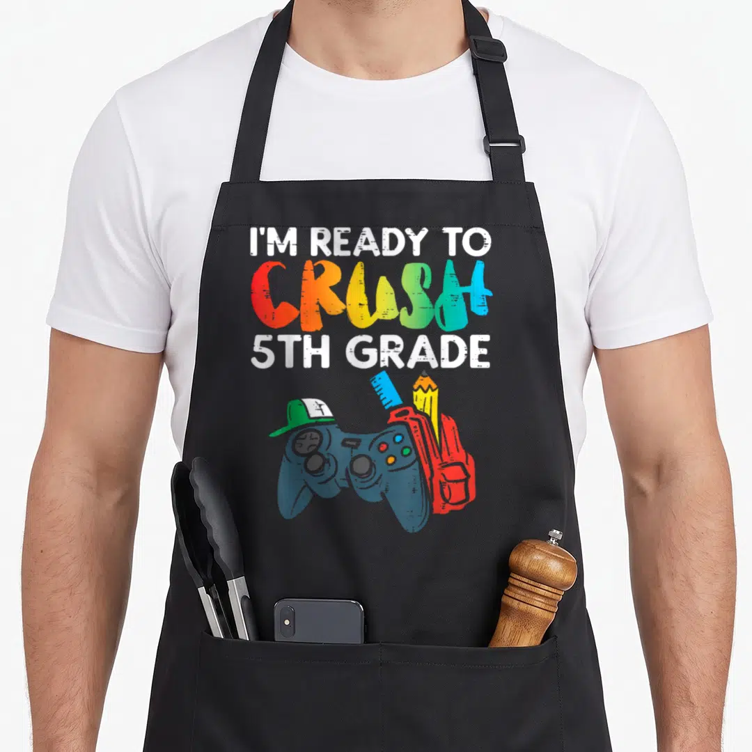 Crush ready grade apron - Rainbow text I'M READY TO CRUSH 5TH GRADE above game controller, red backpack, pencil, ruler — shown on a black cotton canvas apron against a white background