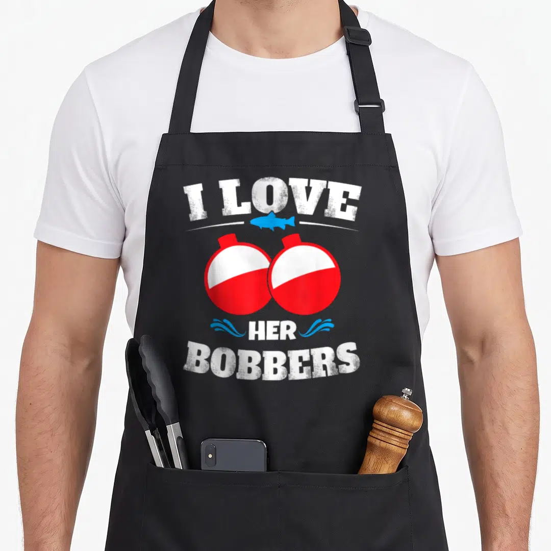 Bobber fishing apron - I Love Her Bobbers text with two red white bobbers and blue fish illustration — shown on a black cotton canvas apron against a white background