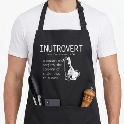 Inutrovert dog apron - White INUTROVERT definition text next to sitting shiba inu silhouette — shown on a black cotton canvas apron against a white background